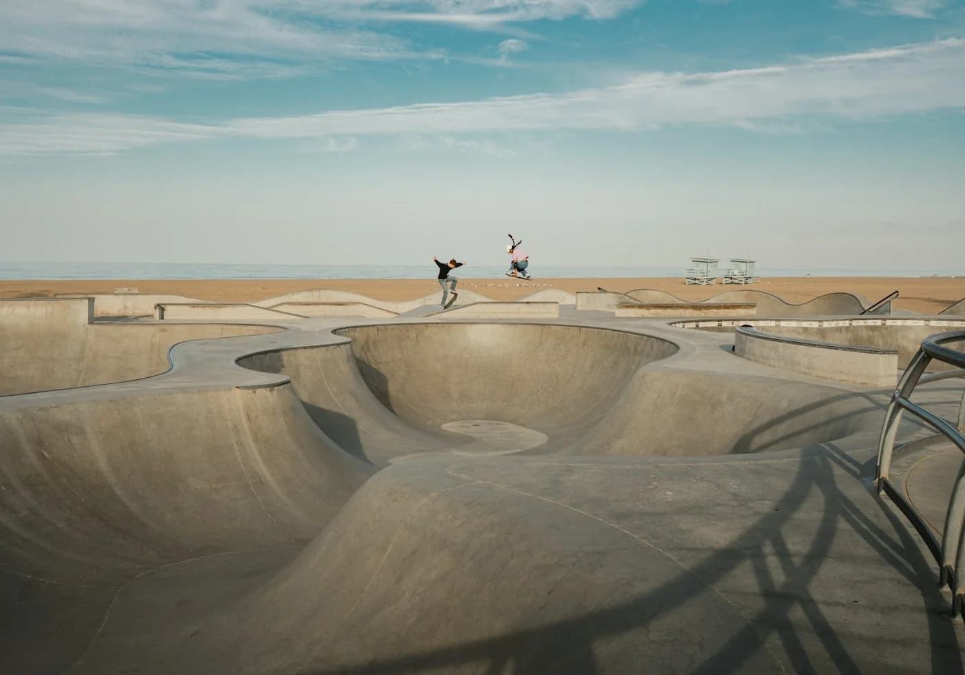 Two skateboarders ride the concrete bowls at Venice Beach Skatepark beside the Pacific Ocean.